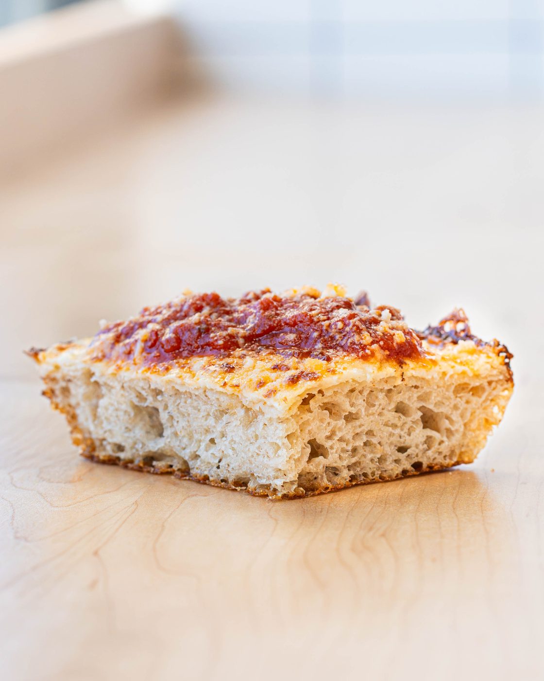 A square slice seen from the side on a wooden board, showing the airy crust, browned cheese edge, and sauce on top